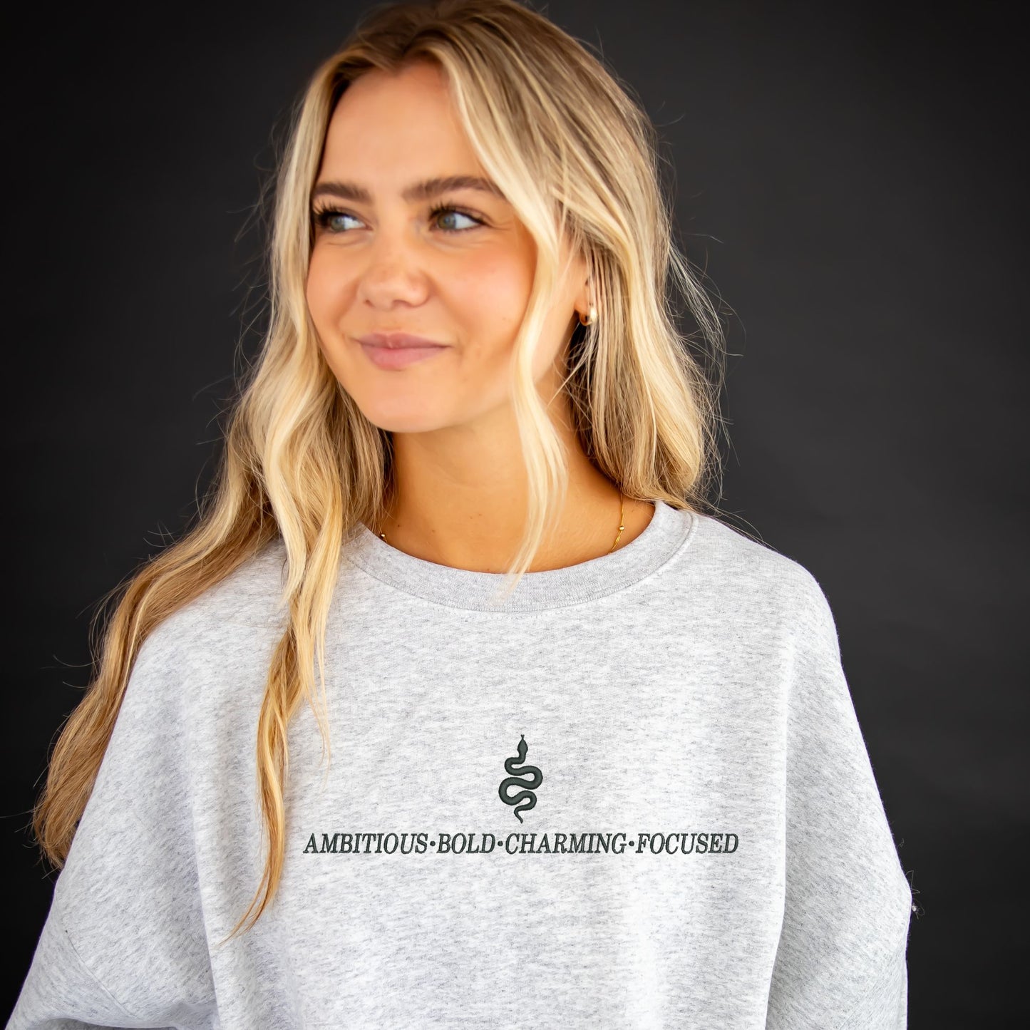 Girl wearing an ash crewneck sweatshirt with custom embroidered Slytherin snake and text reading ambitious, bold, charming, focused.