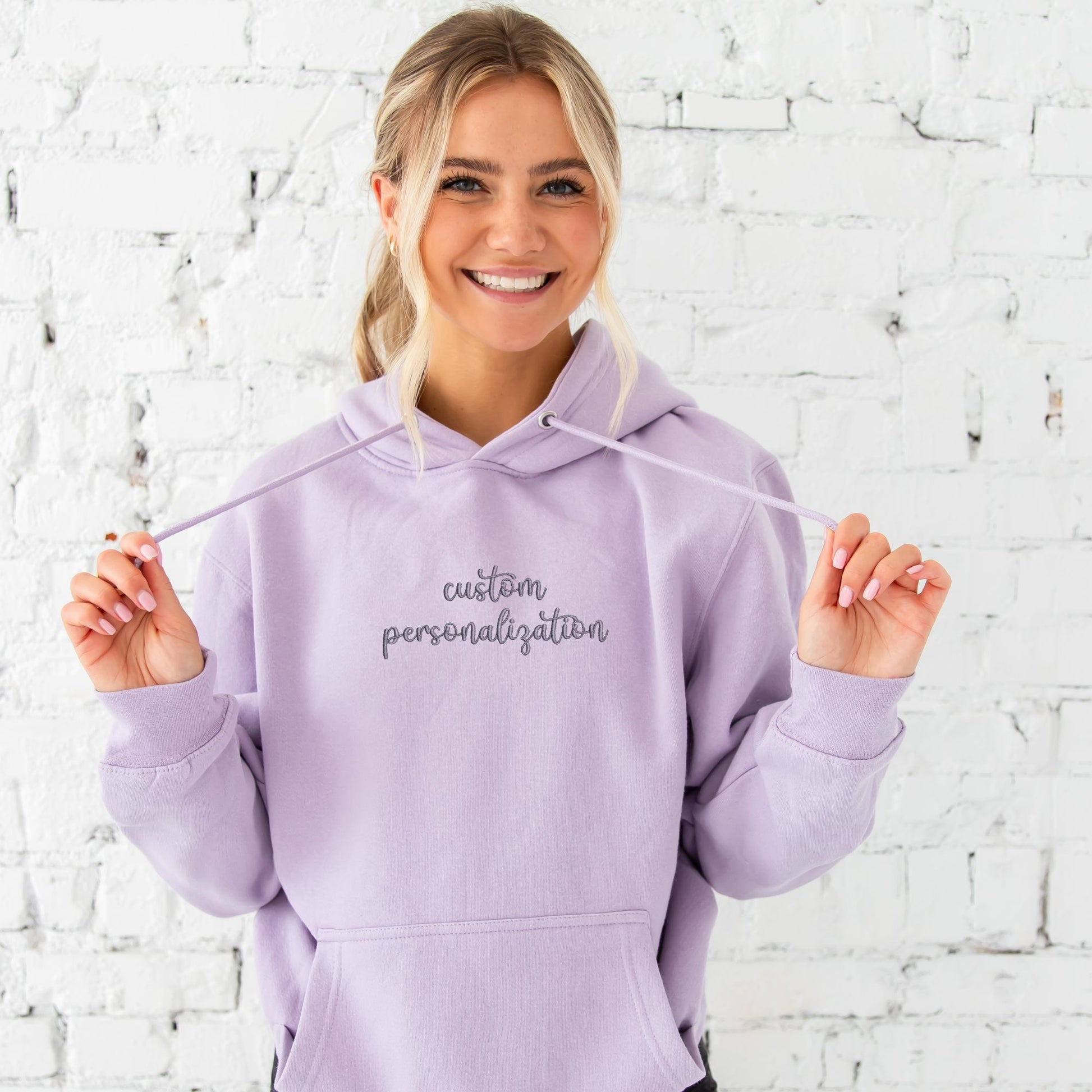 young woman wearing a lilac hoodie with custom personalization in smoky orchid thread embroidered across the chest