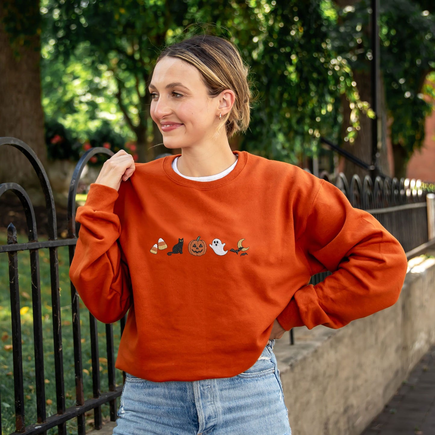woman wearing a texas orange crewneck sweatshirt with halloween icons embroidered as following: candy corn, black cat, jack-o-lantern, ghost, and bats flying over the moon.