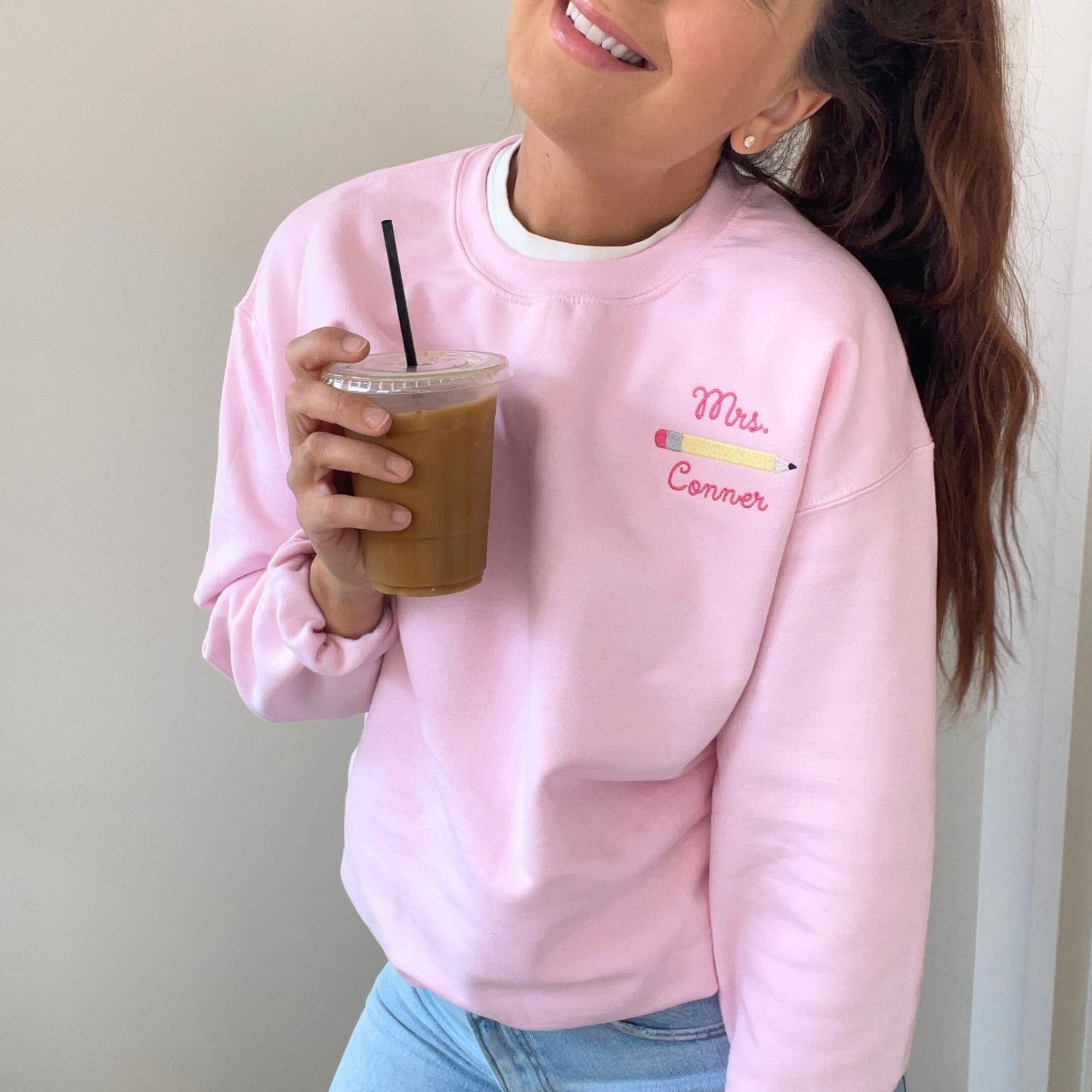 woman wearing a pink crewneck sweatshirt with 'Mrs. Conner' and pencil embroidered design on the left chest, she's holding an iced coffee