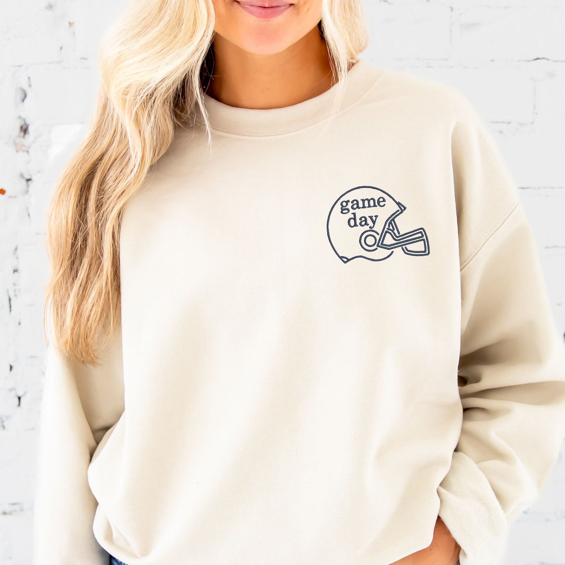 woman wearing a sand crewneck with embroidered game day and football helmet on the left chest in navy thread