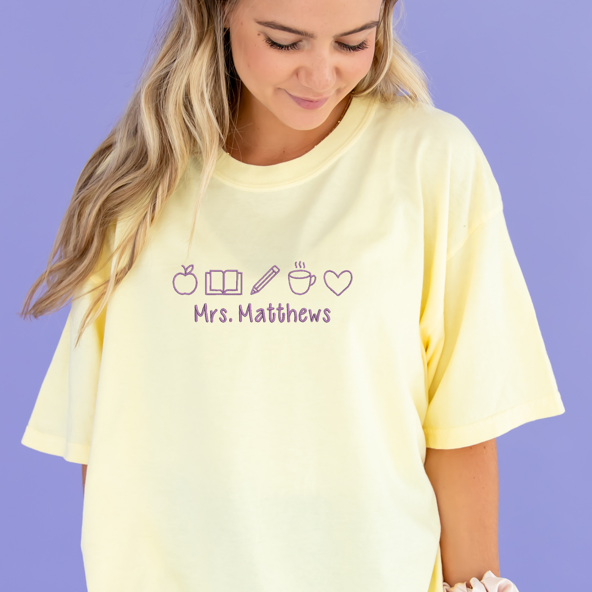 young woman wearing a banana yellow comfort colors t-shirt with embroidered teacher icons in pink thread across the chest and custom text underneath