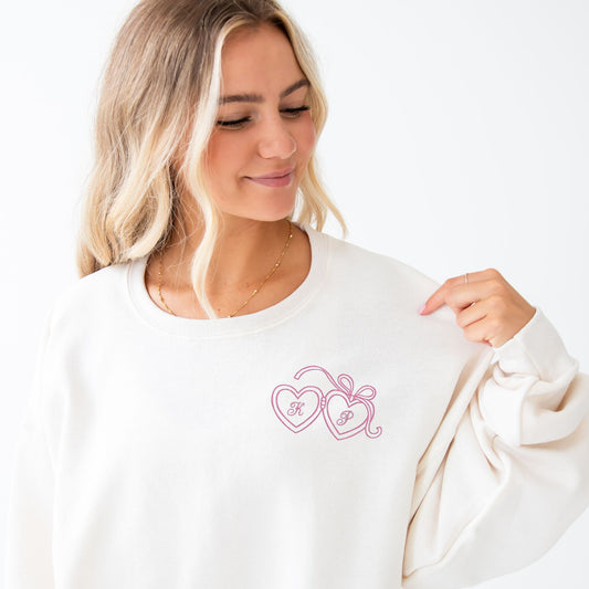 girl wearing a sweet cream crewneck sweatshirt with a design of an outlined teddy bear holding a heart with an initial in the center embroidered on the left chest in pink thread