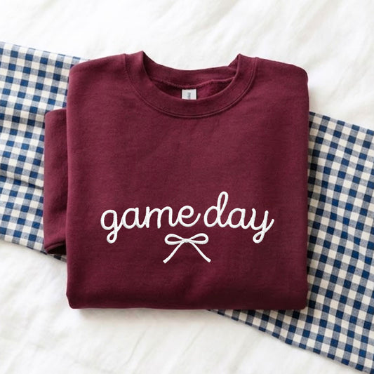 A folded maroon crewneck sweatshirt with 'game day' and a bow embroidered design in white on the front, displayed on a white textured blanket with blue-and-white gingham fabric underneath.