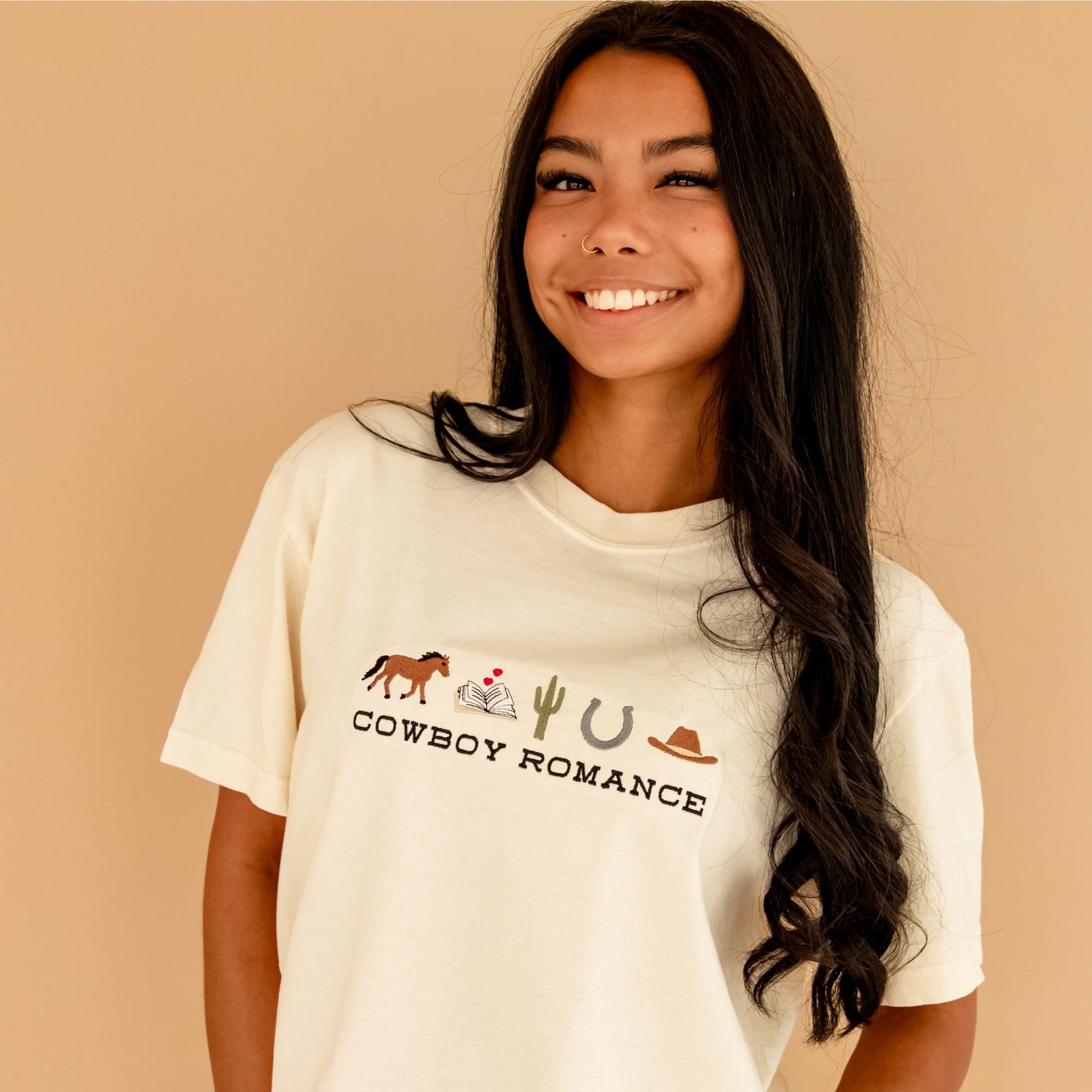 A brunette woman standing in front of a camel colored backdrop wearing an ivory comfort colors t-shirt with a custom embroidered design on the center chest featuring "cowboy romance" in a western inspired font centered underneath the following western icon: horse, book with a mini heart above, cactus, horseshoe, and cowboy hat.