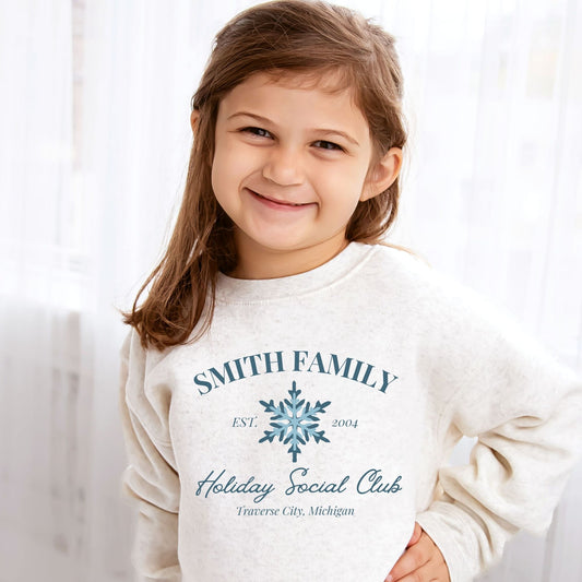 Little girl wearing a natural heather crewneck with a custom print featuring a family name, Est date, snowflake, holiday social club, and location in blue