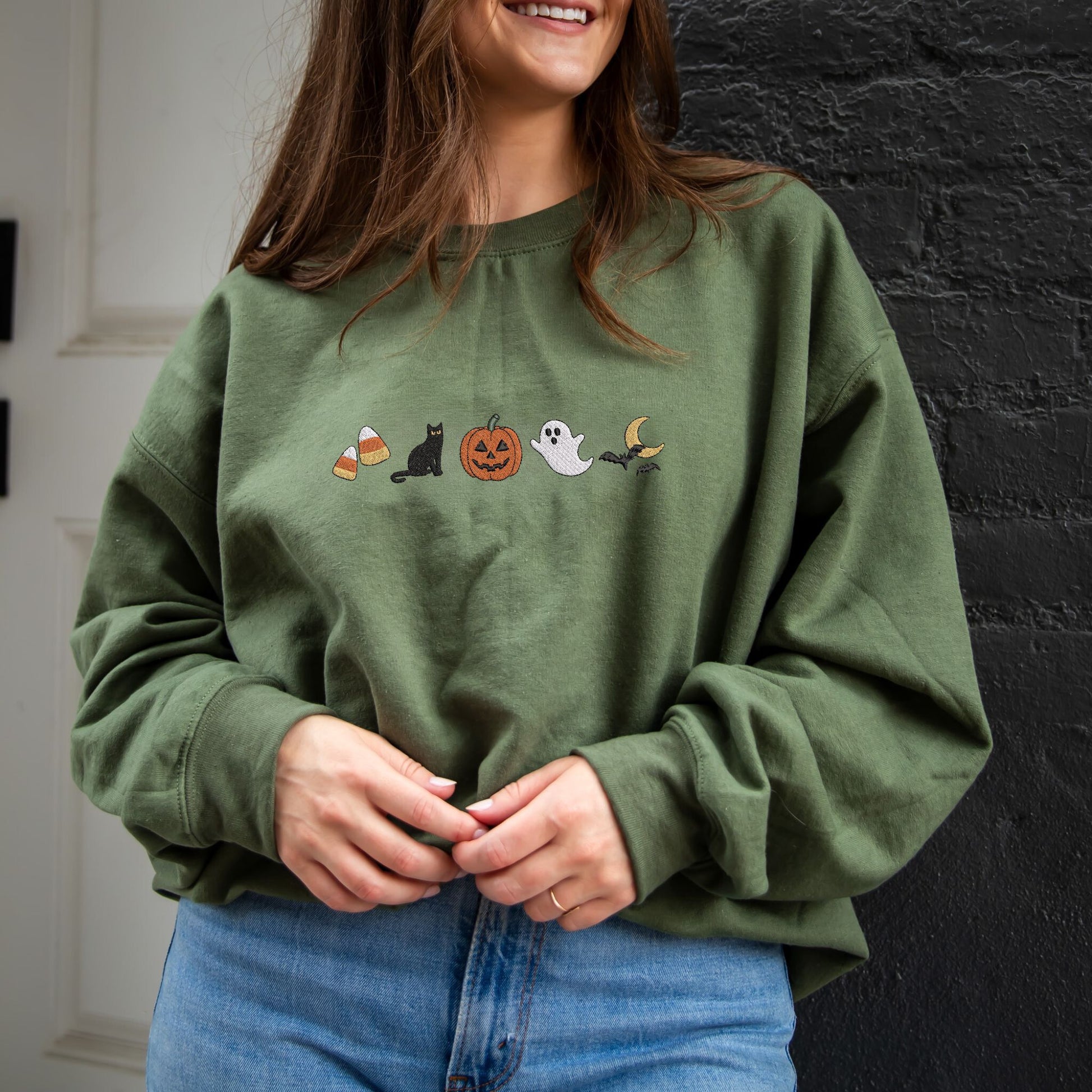 woman wearing a military green crewneck sweatshirt with halloween icons embroidered as following: candy corn, black cat, jack-o-lantern, ghost, and bats flying over the moon.