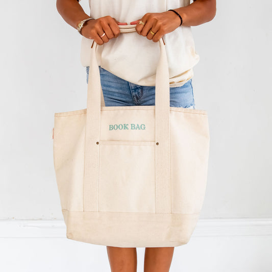 Person holding a canvas tote bag with 'BOOK BAG' embroidered in eucalyptus thread above the front pocket. The person is wearing a cream t-shirt, denim shorts, and a bracelet, standing against a white background. The natural canvas tote features sturdy handles and a minimalist design.