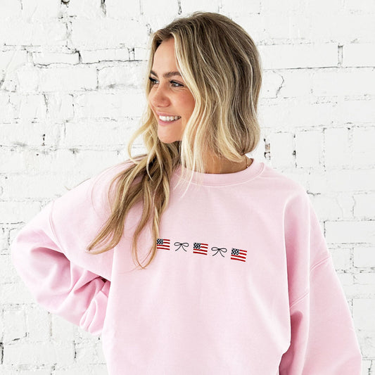 girl with long blonde hair standing in front of a white brick wall, and she's wearing an oversized light pink crewneck sweatshirt with an embroidered dainty bow and mini USA flag alternating pattern embroidered design