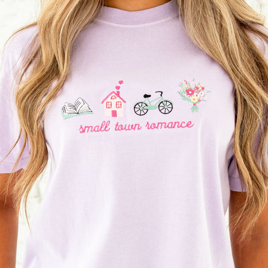 girl standing in front of a white brick wall and she's wearing a cropped orchid comfort colors t-shirt with a cute small town romance embroidered icon design on the center chest featuring: an open book, a house with hearts coming out of the chimney, a vintage bike, and a bouquet of colorful flowers.