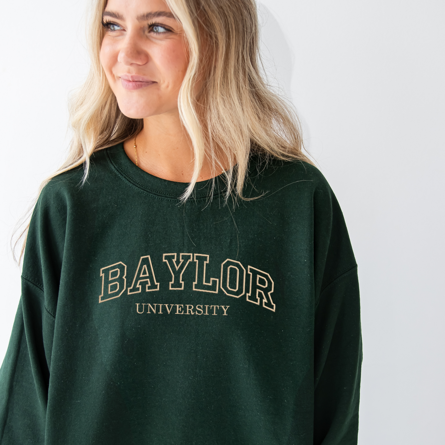 young woman wearing a forest green crewneck sweatshirt with embroidered Baylor University design in camel thread across the chest