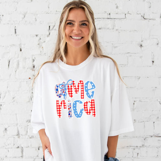 a woman standing in front of a white brick wall wearing a white comfort colors t-shirt with coquette bow lace America floral and gingham print on the center chest