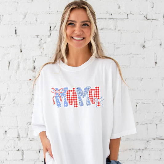 a woman standing in front of a white brick wall wearing a white comfort colors t-shirt with coquette bow lace mama floral and gingham print on the center chest
