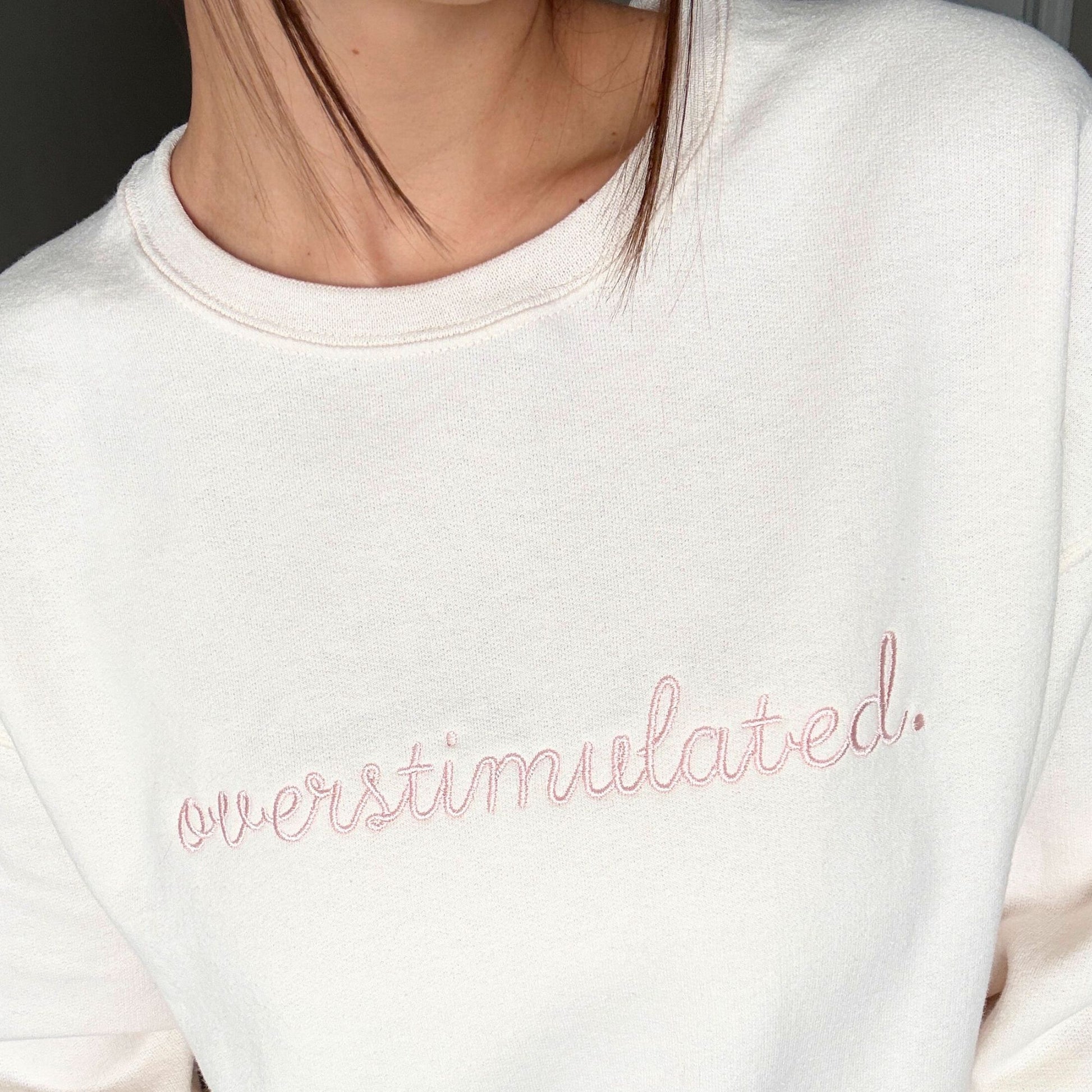 woman wearing a sweet cream crewneck sweatshirt with custom overstimulated embroidered design in a script font and powder pink thread.
