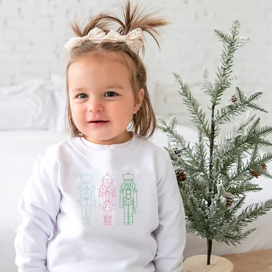 toddler girl wearing a white crewneck sweatshirt with embroidered stitched nutcrackers across the chest in sky blue, pink, and kelly green