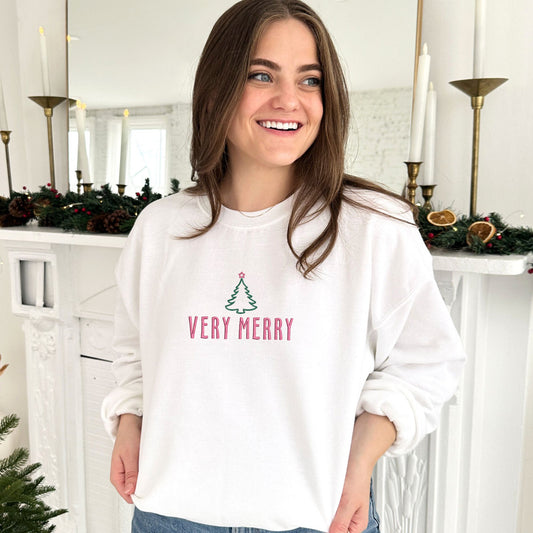 young woman wearing a white crewneck sweatshirt with custom very merry outlined Christmas tree embroidered design in Kelly green and pink