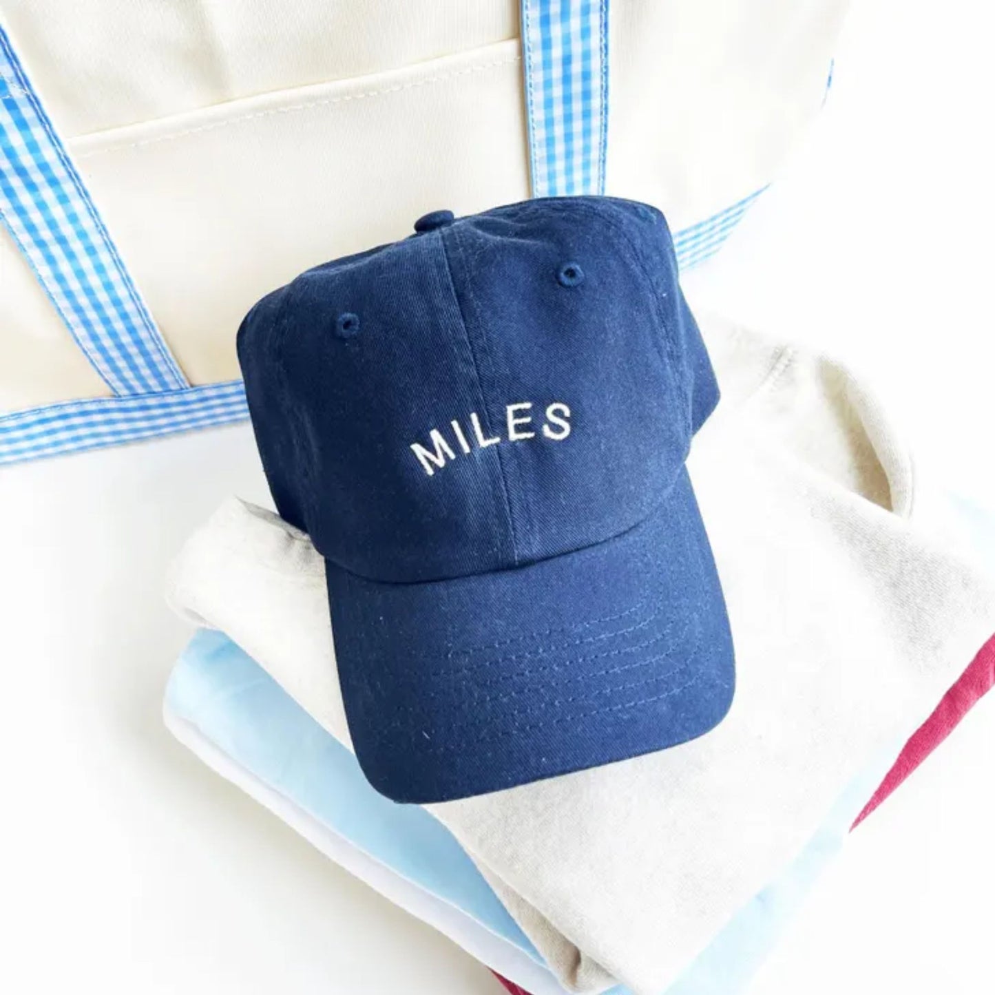 May include: A navy blue baseball cap with a white embroidered word 'MILES' on the front. The cap is sitting on top of a stack of folded clothing in white, light blue, and red.