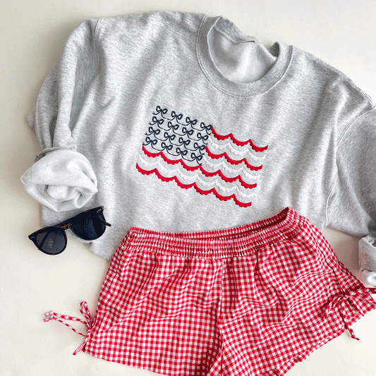 A gray sweatshirt with a red, white, and blue embroidered American flag design. The flag is made of small bows. The sweatshirt is paired with red and white gingham shorts.
