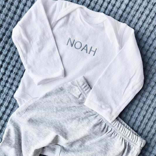 white infant long sleeve bodysuit with custom name embroidered in powder blue thread in Arial uppercase font across the center chest paired with heather grey joggers