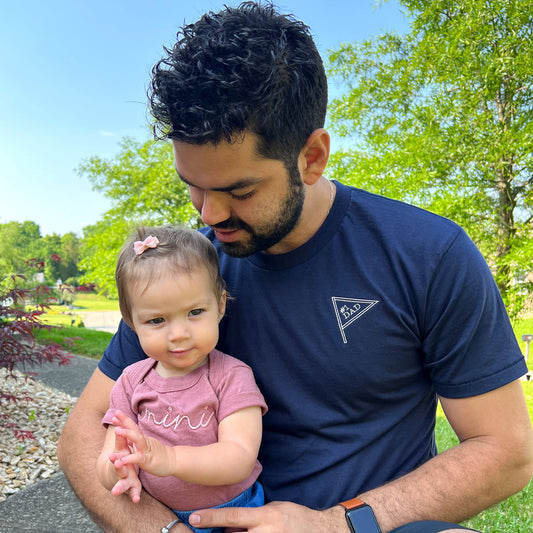 a young dad holding his baby daughter. The dad is wearing a navy crewneck tee with small embroiered pennant on the left chest that reads #1 dad
