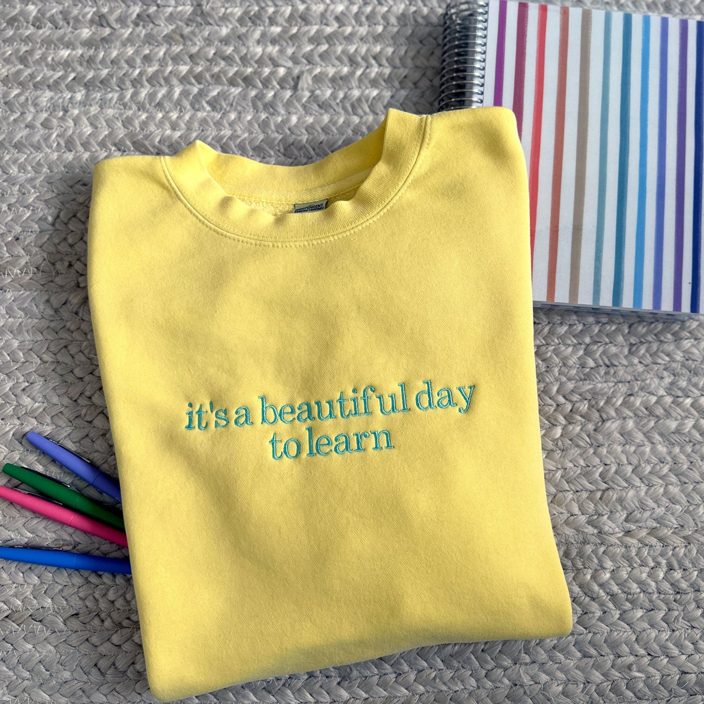 YELLOW SWEATSHIRT WITH 'IT'S A BEAUTIFUL DAY TO LEARN' EMBROIDERED DESIGN ACROSS THE FRONT WITH COLORFUL PENS AND PLANNERS AROUND IT