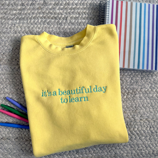 YELLOW SWEATSHIRT WITH 'IT'S A BEAUTIFUL DAY TO LEARN' EMBROIDERED DESIGN ACROSS THE FRONT WITH COLORFUL PENS AND PLANNERS AROUND IT