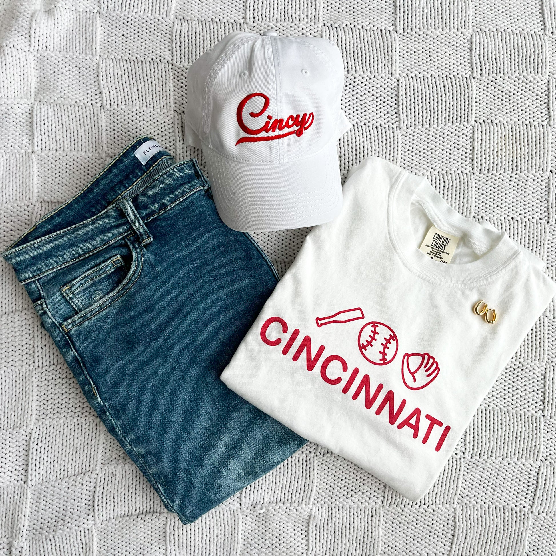 flat lay of jeans, a ehitr baseball icons print tee and an embroidered white  hat with cincy in red