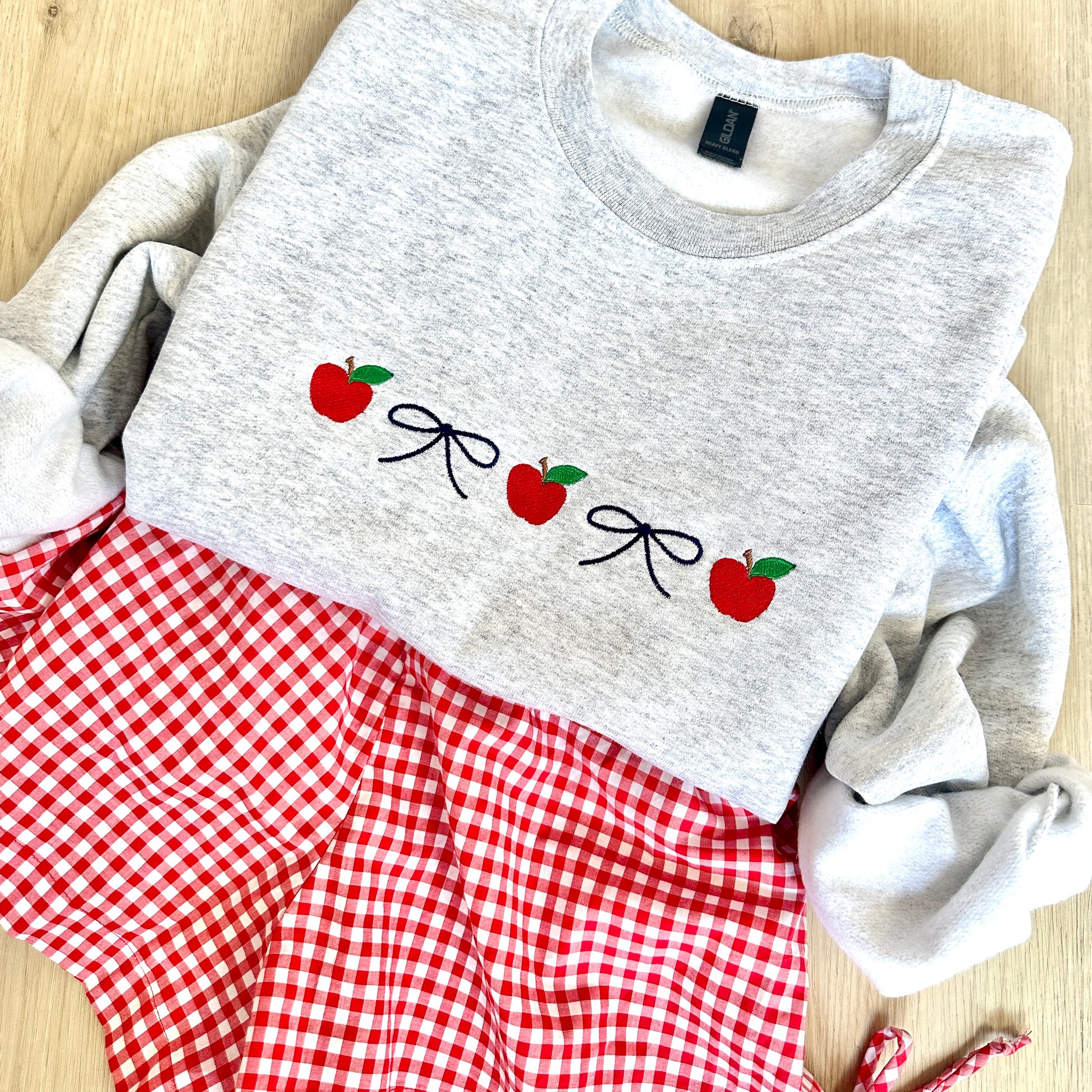 ash crewneck sweatshirt with embroidered navy dainty bows and red apples in an alternating pattern. Sweatshirt is paired with red gingham shorts