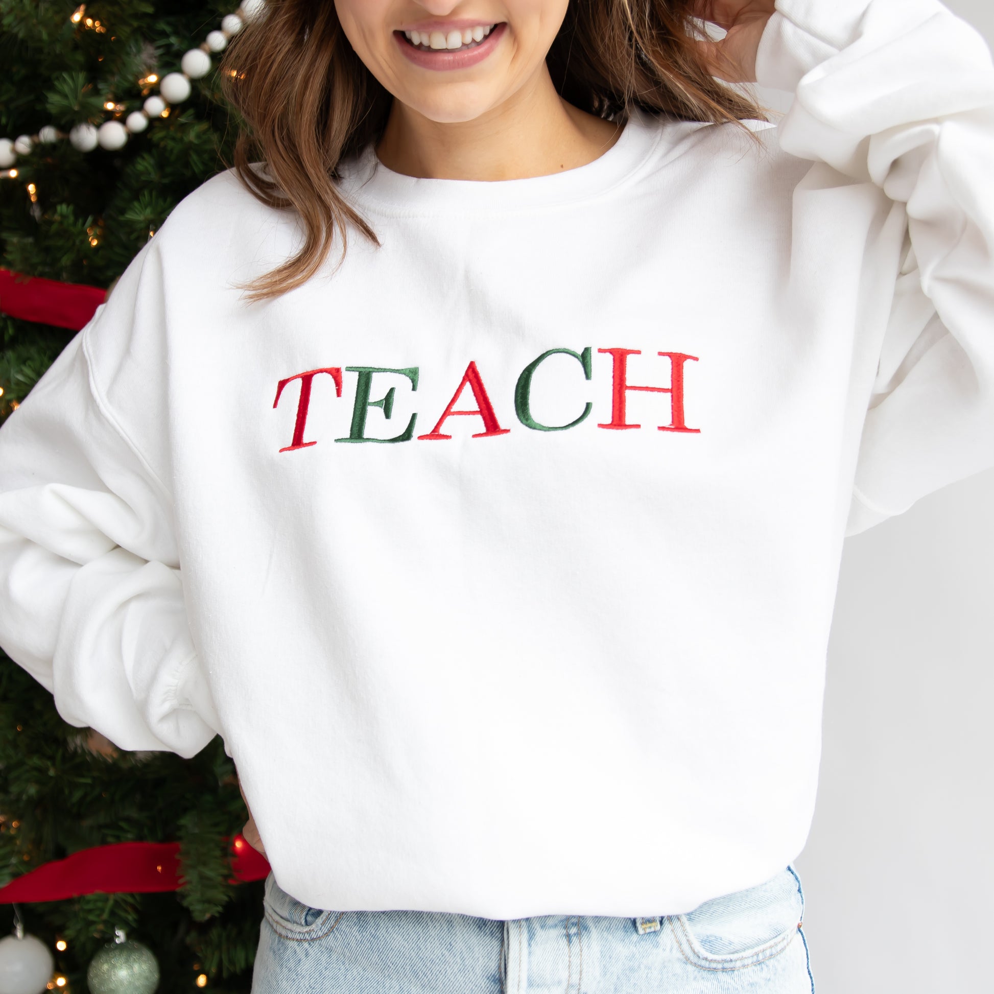 Woman modeling a white crewneck sweatshirt showcasing the word Teach embroidered in red and green Christmas colors.