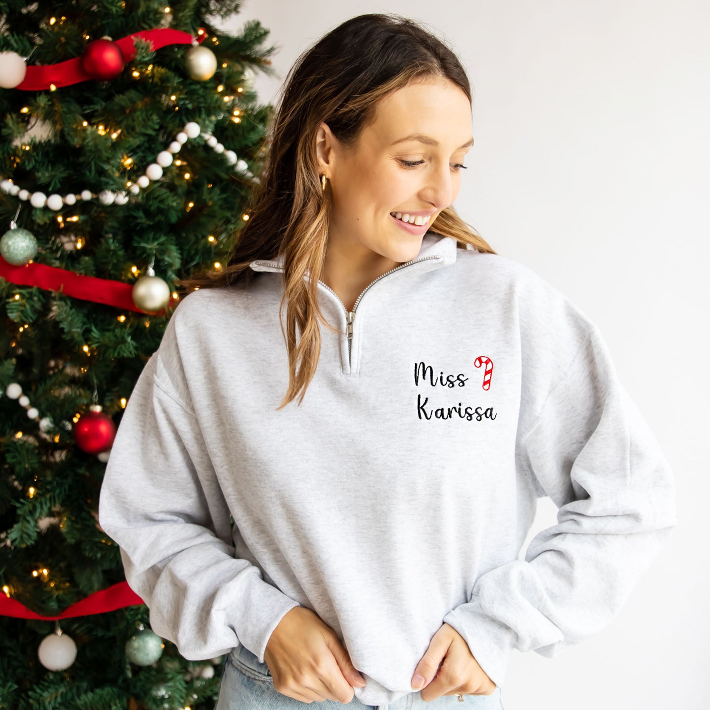 woman standing next to christmas tree wearing a pullover ash quarter zip with custom name and candy cane embroidered design