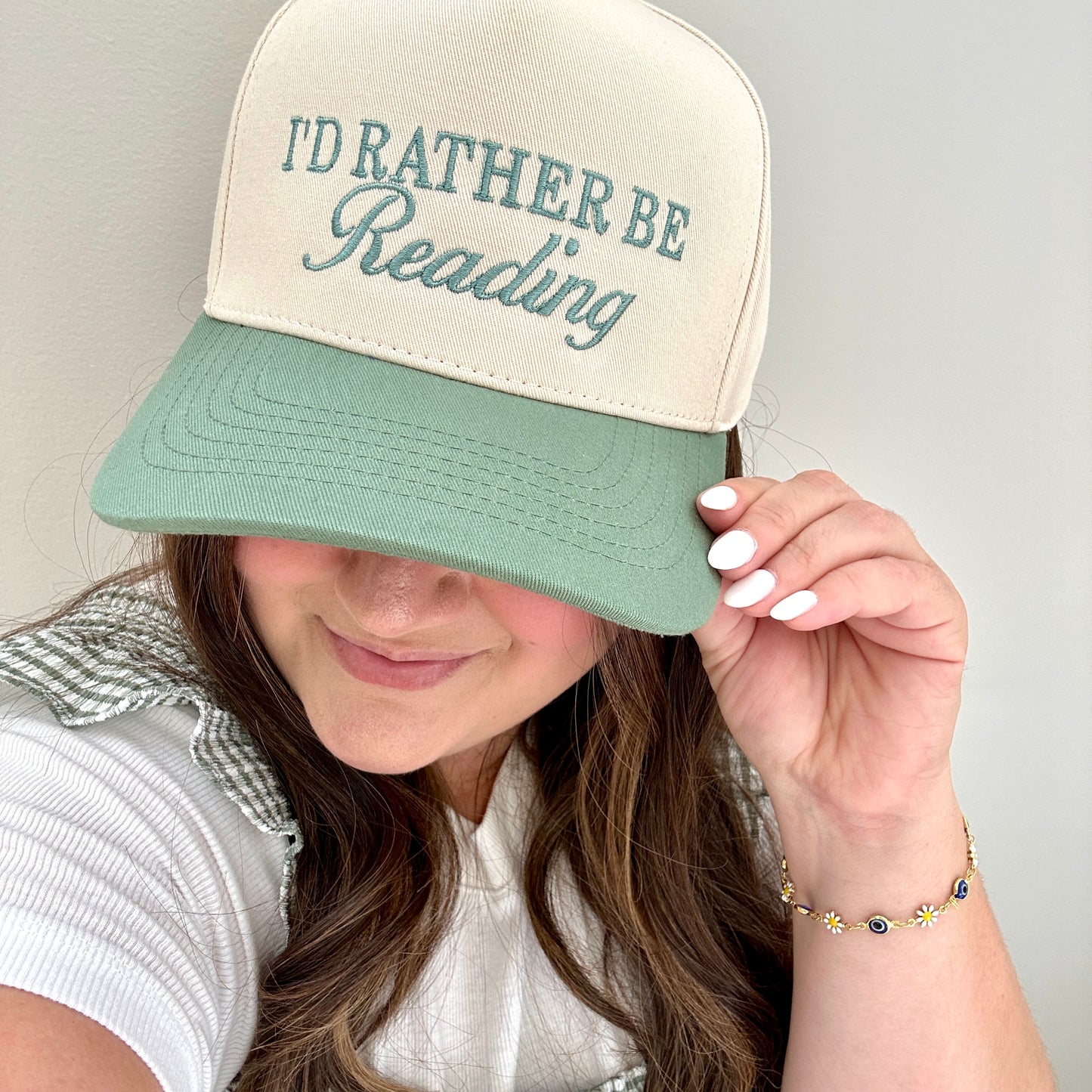 brunette woman wearing and holding the brim of a a two toned canvas-sage trucker hat with custom I'd Rather Be Reading embroidered design in a mixed text design in eucalyptus green thread. She's wearing a gingham green and white dress layered over a white tee and a bracelet with mini daisies and blue beads on a gold chain