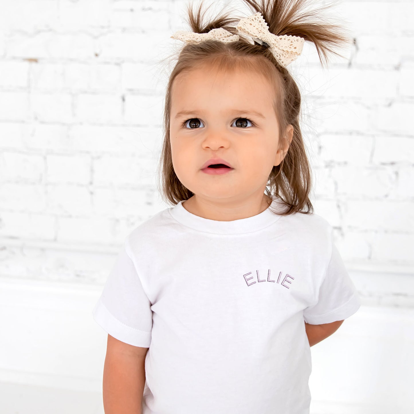 little girl wearing a white tee with her name embroidered on the left chest in a lilac arched font
