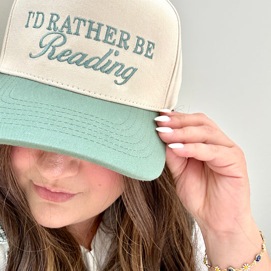 brunette woman wearing and holding the brim of a a two toned canvas-sage trucker hat with custom I'd Rather Be Reading embroidered design in a mixed text design in eucalyptus green thread.