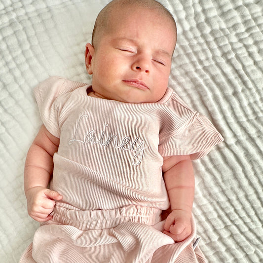 baby girl laying on a muslin towel, and she's wearing a matching pink set featuring a ruffle short sleeve ribbed bodysuit and pleated shorts. Her name is embroidered in a cursive script font across the chest in white thread.