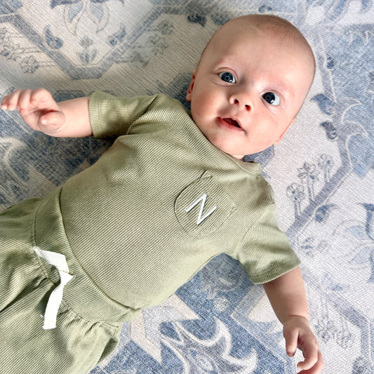 baby boy laying on a blue rug wearing a matching olive green bodysuit and matching pull-on shorts. The bodysuit has a small pocket with a custom initial embroidered on it in white thread.