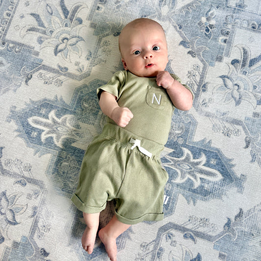 baby boy laying on a blue rug wearing a matching olive green bodysuit and matching pull-on shorts. The bodysuit has a small pocket with a custom initial embroidered on it in white thread.