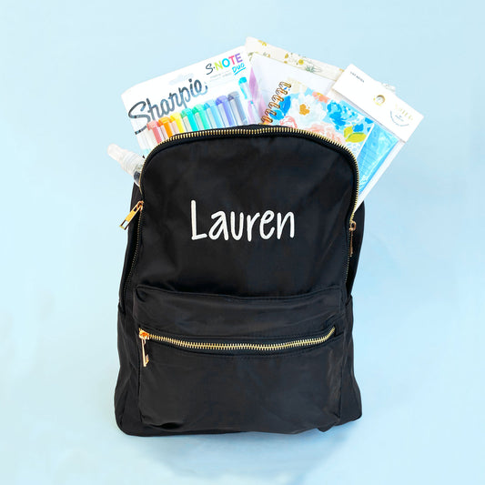 black backpack with embroidered name in white thread cross the front styled with school supplies out the top of the bag