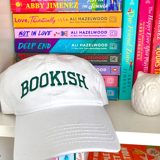white baseball hat with bookish embroidered in athletic font and hunter green thread sitting on a book shelf with colorful books behind it