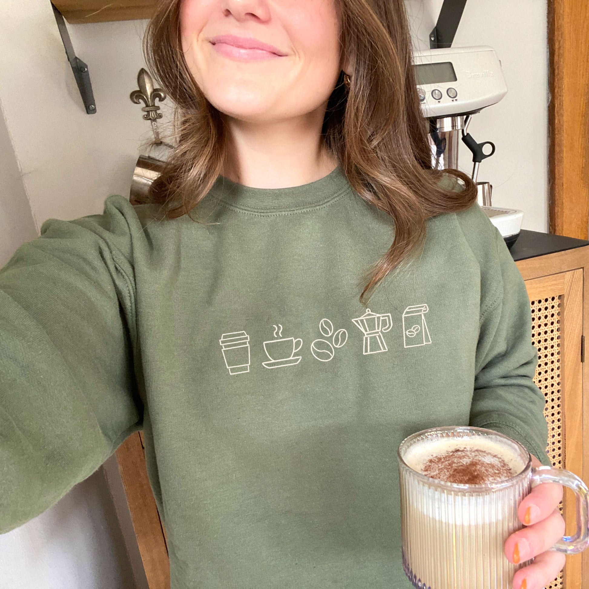 woman wearing a military green crewneck sweatshirt with embroidered coffee icons across the chest in natural thread