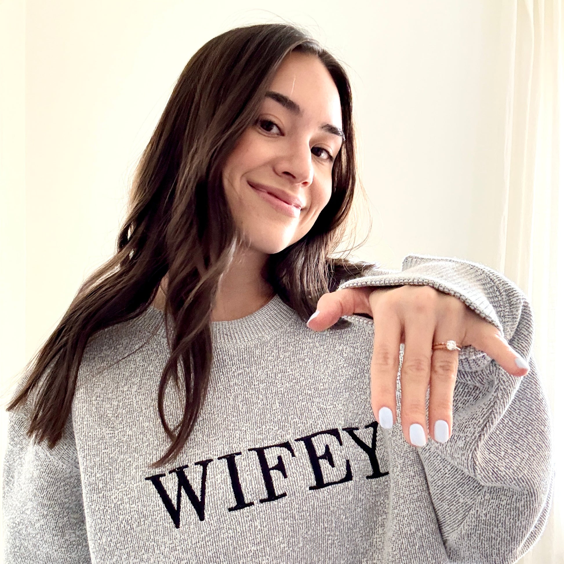 woman showing her wedding ring wearing a cozy salt and pepper knit crew with wifey embroidered across the chest in black bold letters