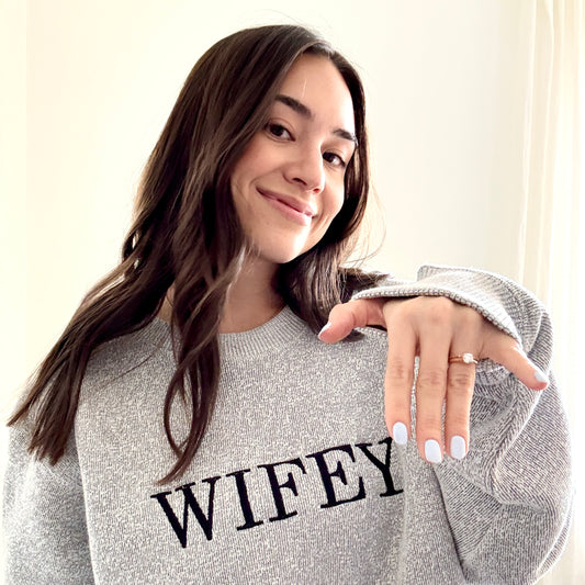 woman showing her wedding ring wearing a cozy salt and pepper knit crew with wifey embroidered across the chest in black bold letters