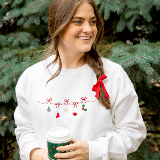 Woman wearing an embroidered white crewneck sweatshirt that features embroidered Christmas garland across the chest.
