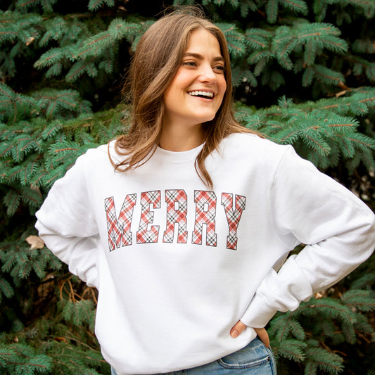 woman standing outside near evergreen tree wearing a white crewneck sweatshirt with MERRY printed in a christmas tartan plaid across the chest