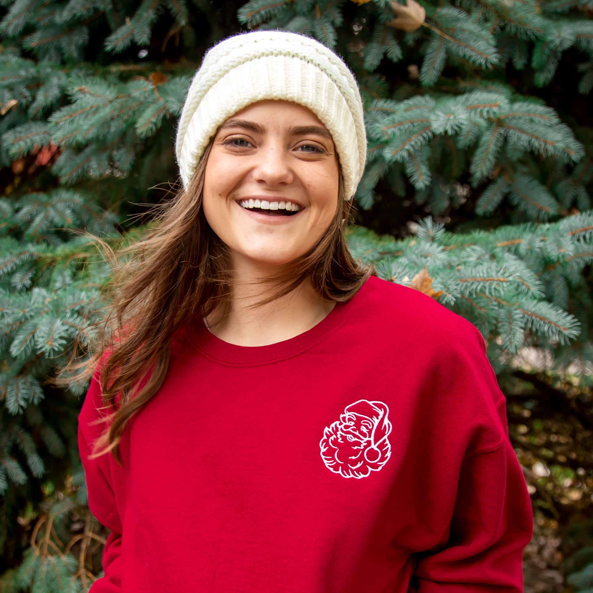 girl outside by a blue spruce tree wearing a red sweatshirt with a santa outline embroidered on the left chest