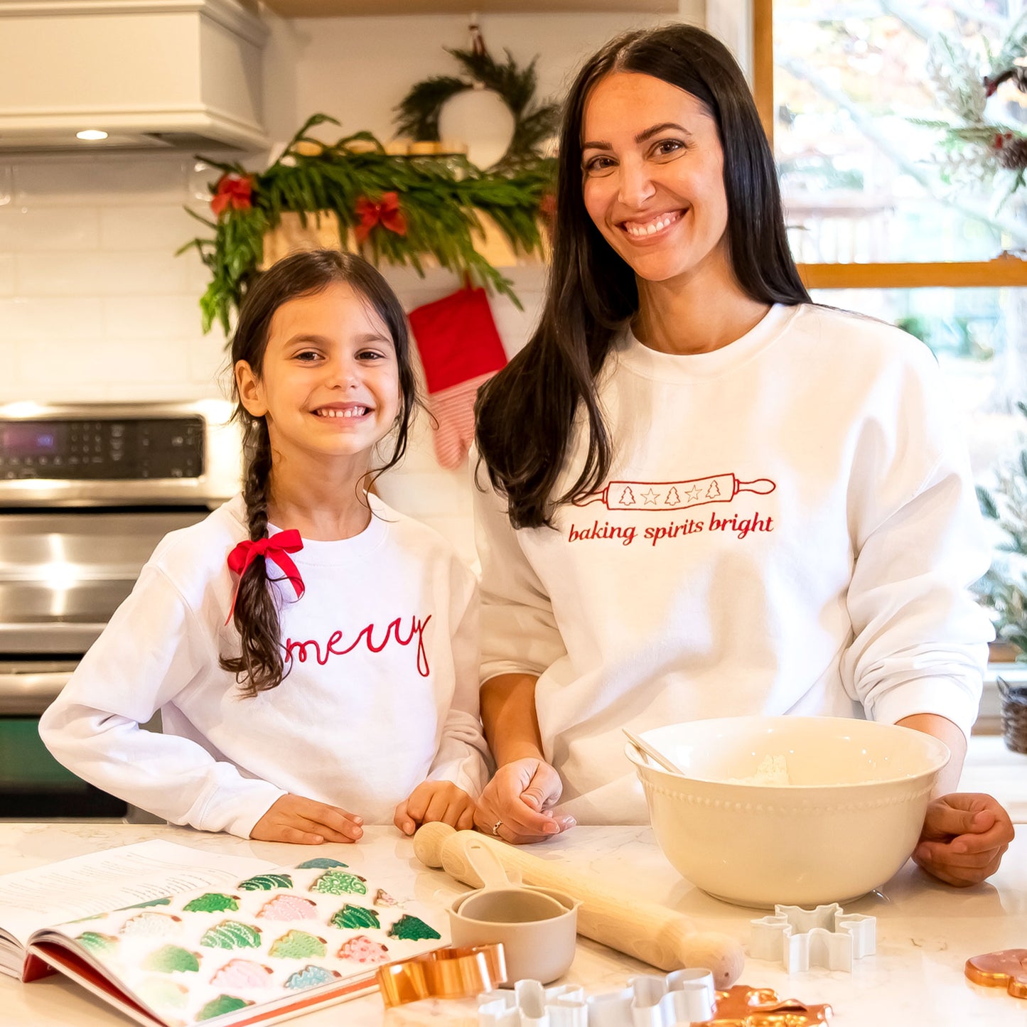 mother and daughter standing in a kitchen baking cookies wearing holiday inspired embroidered sweatshirts