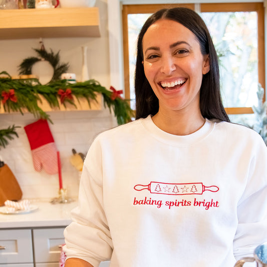 a woman wearing a white pullover sweatshirt with baking spirits bright embroidered across the chest standing in a kitchen deocrated for christmas