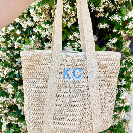 woman holding out a straw knit tote bag in front of a floral bush. tote features a custom embroidery of 2 shadow block initials in a hydrangea blue and powder blue thread