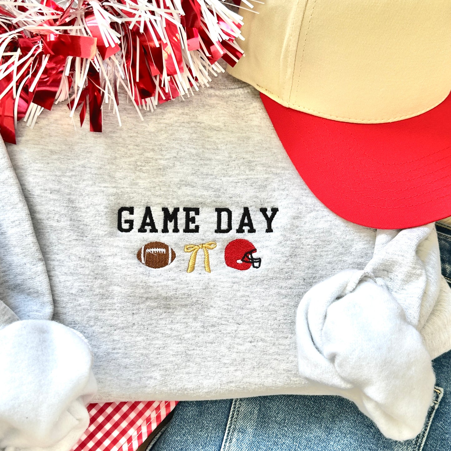 Gray sweatshirt with 'GAME DAY' text and sports icons, surrounded by red and white pom-poms and a red cap.