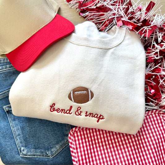 sweet cream sweatshirt with an embroidered football icon and "bend & snap" in a script font, paired with a red cap, jeans, and pom-poms