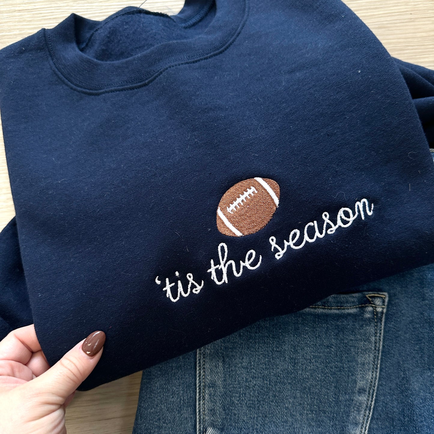 navy blue sweatshirt with a football and script font 'tis the season embroidered design on the center chest paired with blue jeans